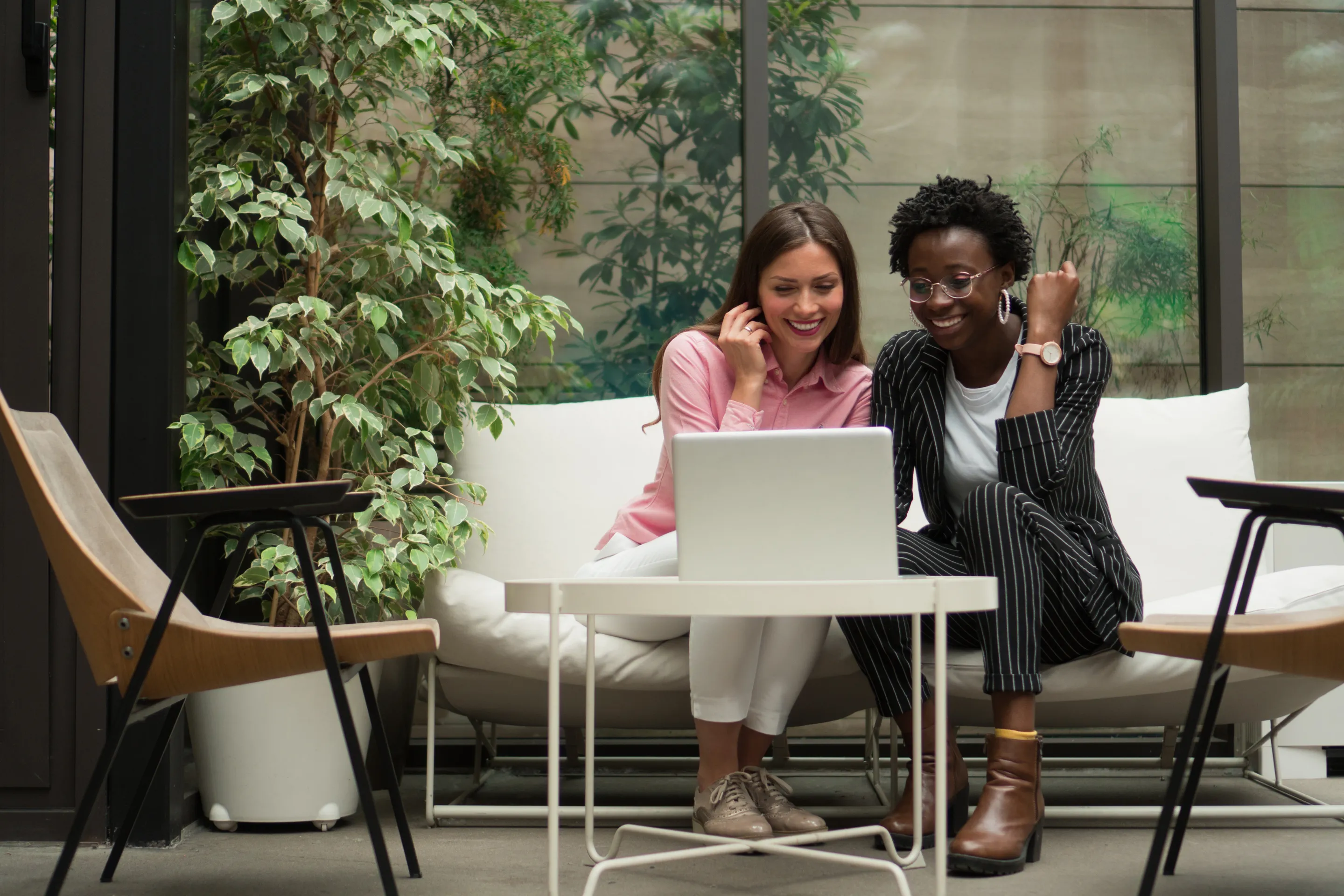 Two colleagues smiling at a laptop on a sofa among plants